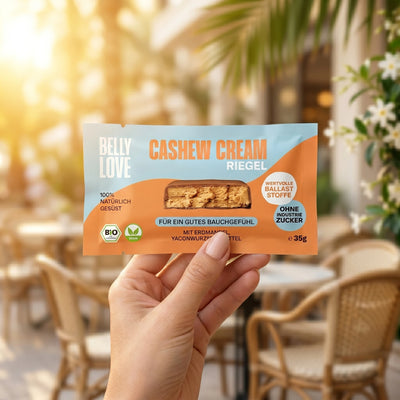 Belly Love Cashew Cream Riegel – Frauenhand hält Verpackung im Café, Bio-Riegel ohne Industriezucker